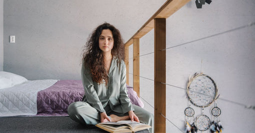 Woman in silk pyjamas reading book by bed in cosy winter evening — July slow evening rituals silk Calliope Studio Australia